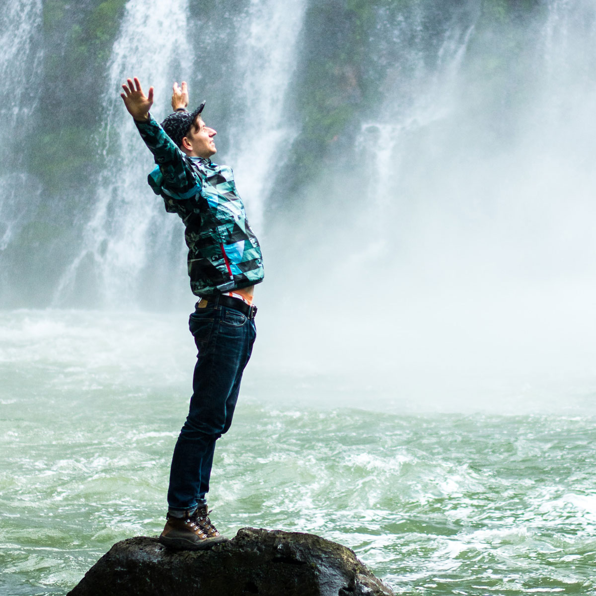 Programm Atem Person steht mit erhobenen Armen auf einem Felsen vor einem Wasserfall