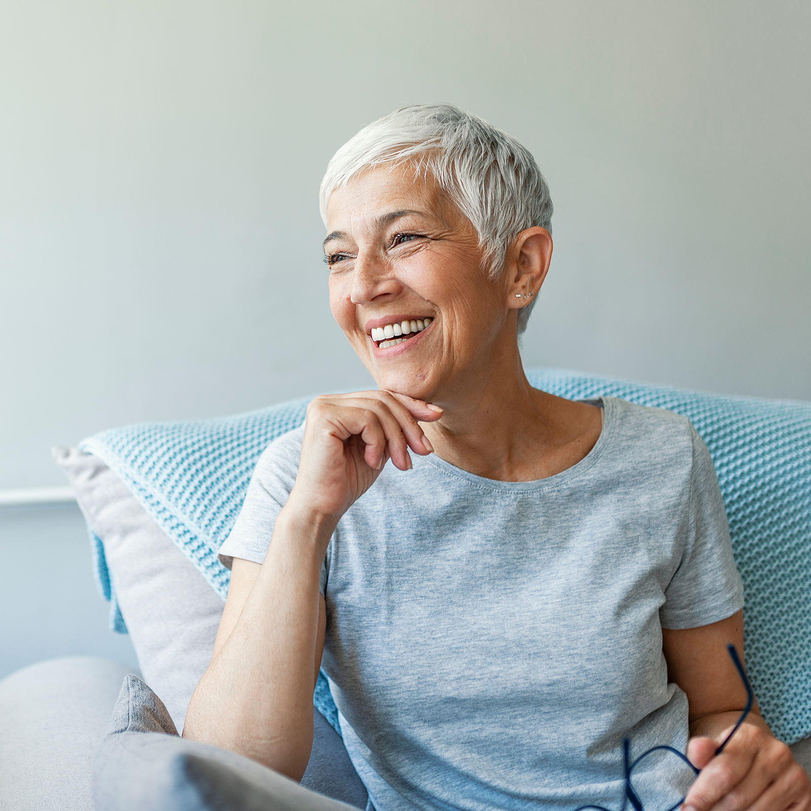 Programm Covidcare Ältere Person mit kurzen grauen Haaren sitzt nachdenklich auf einem Sofa, trägt graues T-Shirt und hält eine Brille in der Hand